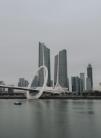 A modern architectural scene featuring a futuristic bridge with a large circular design element spanning across calm water. The backdrop includes several tall skyscrapers constructed with glass and steel, reflecting a modern urban landscape. The sky above is overcast, contributing to a serene and somewhat subdued atmosphere.