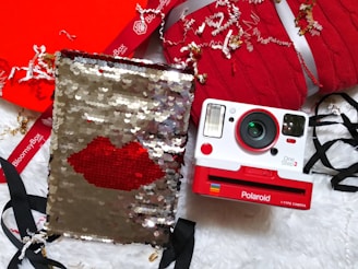 A Polaroid camera with a red and white design is positioned on a fluffy white surface. Next to it lies a silver sequined pouch with a red lips design. Surrounding them are red knit fabric, black ribbons, and decorative shredded paper.