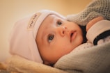A baby with wide eyes gazes upward, wearing a white hat with a label containing text. The baby is wrapped in a soft, textured blanket, with a gentle expression and rosy cheeks.