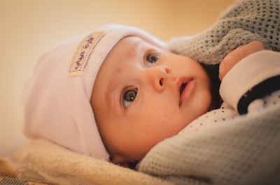 A baby with wide eyes gazes upward, wearing a white hat with a label containing text. The baby is wrapped in a soft, textured blanket, with a gentle expression and rosy cheeks.