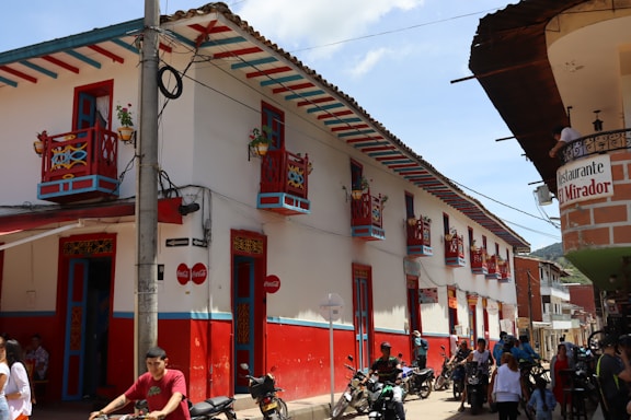 A vibrant street scene with a colonial-style building featuring colorful red and blue accents, decorative balconies with potted plants, and signs for a restaurant named 'El Mirador'. People and motorcycles are present on the street in a bright, sunny atmosphere.