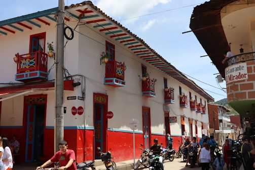 A vibrant street scene with a colonial-style building featuring colorful red and blue accents, decorative balconies with potted plants, and signs for a restaurant named 'El Mirador'. People and motorcycles are present on the street in a bright, sunny atmosphere.