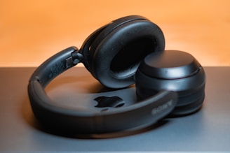 A pair of black over-ear headphones rests on a flat surface with an indistinct logo visible beneath. The background features a soft, warm glow creating a slight bokeh effect.