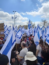 A large crowd gathers outdoors, holding numerous flags with blue and white colors and a prominent star symbol. People of various ages are present, some wearing hats and casual clothing. The sky is partly cloudy, and there is a sense of movement as the flags flutter.