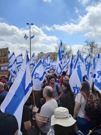 A large crowd gathers outdoors, holding numerous flags with blue and white colors and a prominent star symbol. People of various ages are present, some wearing hats and casual clothing. The sky is partly cloudy, and there is a sense of movement as the flags flutter.
