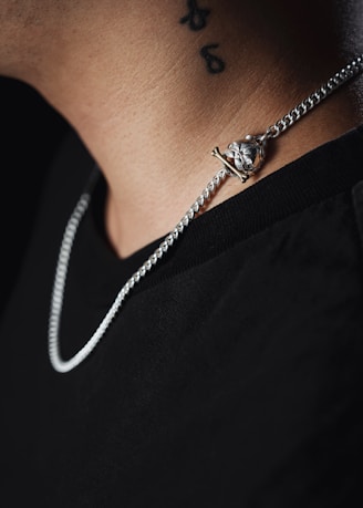 A close-up of a person's neck featuring a silver chain with a pendant shaped like a skull. The person is wearing a black shirt, and there is a black tattoo visible on the neck.
