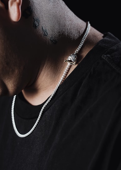 A close-up of a person's neck displaying a silver chain with a unique pendant. The background is dark, highlighting the texture of the chain and skin. The person is wearing a dark shirt, and there are small, dark tattoos visible on the neck.