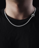 A close-up view of a person's neck wearing a silver chain necklace. The background is dark, and the person is wearing a plain black shirt.