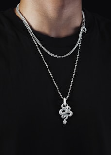 A person is wearing multiple silver chains around their neck against a black background. The most prominent chain features an intricately designed snake pendant with a textured surface.