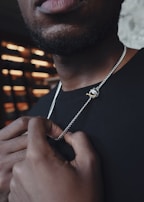 A close-up of a man holding a silver chain with a pendant featuring a small, gold revolver. The focus is on the jewelry and his hands, while the upper part of his face is out of the frame. The lighting creates a contrast against a blurry background composed of horizontal light strips.