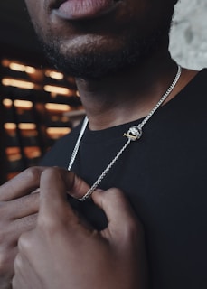 A close-up of a man holding a silver chain with a pendant featuring a small, gold revolver. The focus is on the jewelry and his hands, while the upper part of his face is out of the frame. The lighting creates a contrast against a blurry background composed of horizontal light strips.