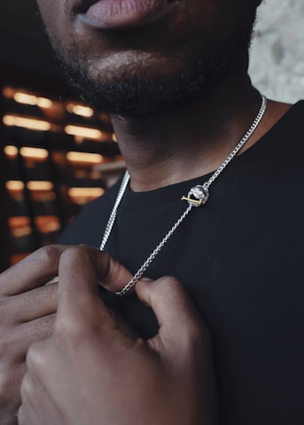 A close-up of a man holding a silver chain with a pendant featuring a small, gold revolver. The focus is on the jewelry and his hands, while the upper part of his face is out of the frame. The lighting creates a contrast against a blurry background composed of horizontal light strips.