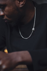 A close-up view of a person wearing a dark long-sleeve shirt and a silver chain necklace. Their face is partially visible and turned sideways, showing an earring. The lighting is dim and creates a moody atmosphere.