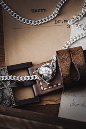 A close-up of a detailed leather strap with silver chains and a metallic skull emblem. The background includes vintage paper documents, partly in German, adding a rustic and historical ambiance.