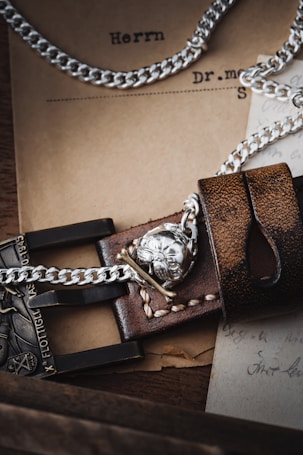 A close-up of a detailed leather strap with silver chains and a metallic skull emblem. The background includes vintage paper documents, partly in German, adding a rustic and historical ambiance.