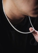 A close-up view of a person's neck wearing a silver chain necklace. The individual is wearing a dark-colored shirt, and the fingers are holding a part of the chain. The focus is on the texture and shine of the chain against the skin.