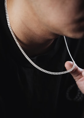 A close-up view of a person's neck wearing a silver chain necklace. The individual is wearing a dark-colored shirt, and the fingers are holding a part of the chain. The focus is on the texture and shine of the chain against the skin.