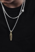 A close-up view of two silver chains with pendants against a plain black background. One pendant is a gold, segmented piece, while the other is a silver heart. The chains have a shiny, polished appearance.