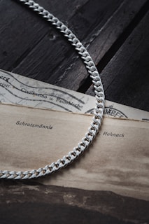 A silver chain with a tightly interlocking link pattern is elegantly placed on top of an old, sepia-toned document. The paper shows visible signs of aging, with printed text featuring words such as 'Schratemännle' and 'Hohnack'. The background surface appears to be a dark, textured wood, enhancing the contrast with the shiny chain.