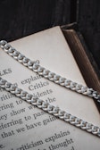 A close-up view of a silver chain resting on an open book. The book's text is partially visible, showcasing words like 'explains,' 'Critics,' and 'incidents.' The chain's metal links are glossy and reflective, creating a contrast against the textured paper pages.