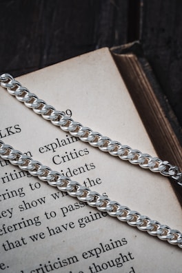 A close-up view of a silver chain resting on an open book. The book's text is partially visible, showcasing words like 'explains,' 'Critics,' and 'incidents.' The chain's metal links are glossy and reflective, creating a contrast against the textured paper pages.