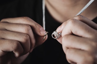 Hands holding a necklace with a ring pendant, closely focused, showcasing the intricate details of the jewelry against a dark background.