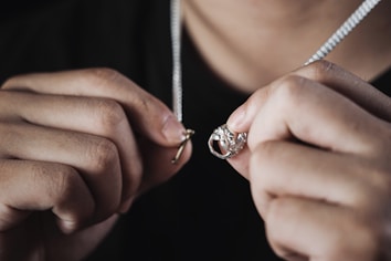 Hands holding a necklace with a ring pendant, closely focused, showcasing the intricate details of the jewelry against a dark background.