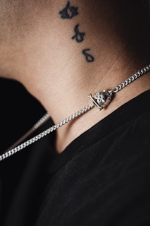 A close-up view of a person's neck and part of their shoulder, showing a silver chain necklace with a pendant resting on the collarbone. There is a tattoo on the neck consisting of small black designs. The person is wearing a dark-colored shirt.