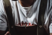 A person wearing a white t-shirt with red text partially visible beneath a pair of dark-colored suspenders. Around their neck, they wear a silver necklace with a cross pendant.