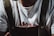 A person wearing a white t-shirt with red text partially visible beneath a pair of dark-colored suspenders. Around their neck, they wear a silver necklace with a cross pendant.