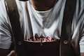 A person wearing a white t-shirt with red text partially visible beneath a pair of dark-colored suspenders. Around their neck, they wear a silver necklace with a cross pendant.
