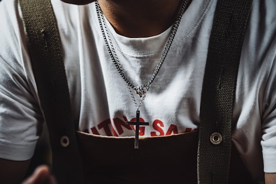 A person wearing a white t-shirt with red text partially visible beneath a pair of dark-colored suspenders. Around their neck, they wear a silver necklace with a cross pendant.