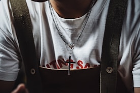 A person wearing a white t-shirt with red text partially visible beneath a pair of dark-colored suspenders. Around their neck, they wear a silver necklace with a cross pendant.