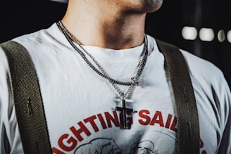 A person is wearing a white T-shirt with red lettering that says 'Fighting Sailor' and features a cartoon graphic. They have layered necklaces, including one with a cross pendant. Green suspenders are also visible, attached to the shoulders of the shirt.