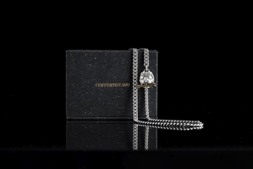 A silver chain with a pendant featuring a lion's head design is elegantly draped over a dark, rectangular box that has a subtle texture. The box bears the text 'COPPERTIST.WU' in a minimalist font. The setting is against a black background, which highlights the metallic shine of the chain and the intricate details of the pendant.