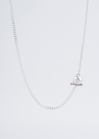 A delicate silver chain necklace with a crystal skull pendant resting on two small crossbones. The background is a light, neutral tone, emphasizing the sleek and minimalist design of the jewelry.