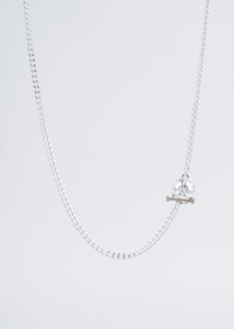 A delicate silver chain necklace with a crystal skull pendant resting on two small crossbones. The background is a light, neutral tone, emphasizing the sleek and minimalist design of the jewelry.