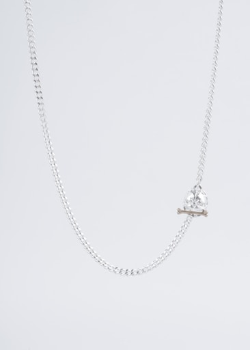 A delicate silver chain necklace with a crystal skull pendant resting on two small crossbones. The background is a light, neutral tone, emphasizing the sleek and minimalist design of the jewelry.