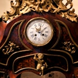 Ornate antique clock with intricate golden details and carved floral decorations. The clock face displays Roman numerals and is encased in a richly varnished wooden frame. A small cherub is featured at the base of the clock.