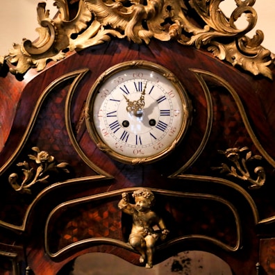 Ornate antique clock with intricate golden details and carved floral decorations. The clock face displays Roman numerals and is encased in a richly varnished wooden frame. A small cherub is featured at the base of the clock.