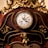 Ornate antique clock with intricate golden details and carved floral decorations. The clock face displays Roman numerals and is encased in a richly varnished wooden frame. A small cherub is featured at the base of the clock.