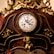 Ornate antique clock with intricate golden details and carved floral decorations. The clock face displays Roman numerals and is encased in a richly varnished wooden frame. A small cherub is featured at the base of the clock.