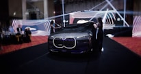 A luxury car unveiling event with two women removing a black cloth from a sleek, illuminated vehicle. The backdrop consists of vibrant, swirling light designs, and several people watch attentively from the side.