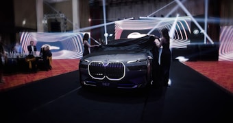 A luxury car unveiling event with two women removing a black cloth from a sleek, illuminated vehicle. The backdrop consists of vibrant, swirling light designs, and several people watch attentively from the side.