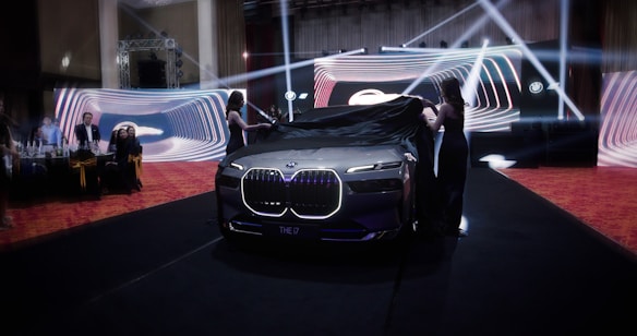 A luxury car unveiling event with two women removing a black cloth from a sleek, illuminated vehicle. The backdrop consists of vibrant, swirling light designs, and several people watch attentively from the side.