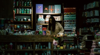 A store display filled with a variety of colorful items such as mugs, gift wrap, toys, and decorative objects. A person, possibly a storekeeper or customer, is standing behind the counter surrounded by shelves. The lighting is dim, giving the shop an intimate and cozy atmosphere.