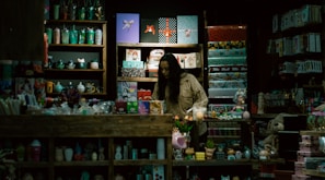 A store display filled with a variety of colorful items such as mugs, gift wrap, toys, and decorative objects. A person, possibly a storekeeper or customer, is standing behind the counter surrounded by shelves. The lighting is dim, giving the shop an intimate and cozy atmosphere.