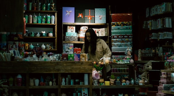 A store display filled with a variety of colorful items such as mugs, gift wrap, toys, and decorative objects. A person, possibly a storekeeper or customer, is standing behind the counter surrounded by shelves. The lighting is dim, giving the shop an intimate and cozy atmosphere.