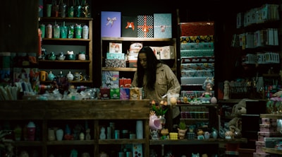 A store display filled with a variety of colorful items such as mugs, gift wrap, toys, and decorative objects. A person, possibly a storekeeper or customer, is standing behind the counter surrounded by shelves. The lighting is dim, giving the shop an intimate and cozy atmosphere.