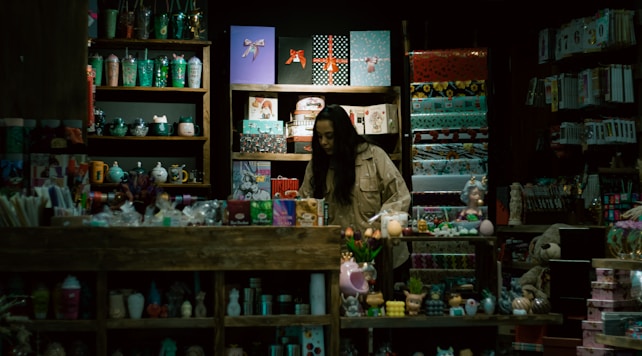 A store display filled with a variety of colorful items such as mugs, gift wrap, toys, and decorative objects. A person, possibly a storekeeper or customer, is standing behind the counter surrounded by shelves. The lighting is dim, giving the shop an intimate and cozy atmosphere.